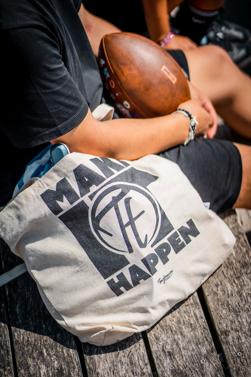 Ein Shopper aus Bio-Baumwolle, naturfarben, mit coolem, schwarzem "Make it happen"-Print, draussen fotografiert, auf einem Holzsteg (Nahaufnahme des Shoppers), im Hintergrund sitzt ein Mann in Tagtraum Kleidung, in der Hand hält er einen Football