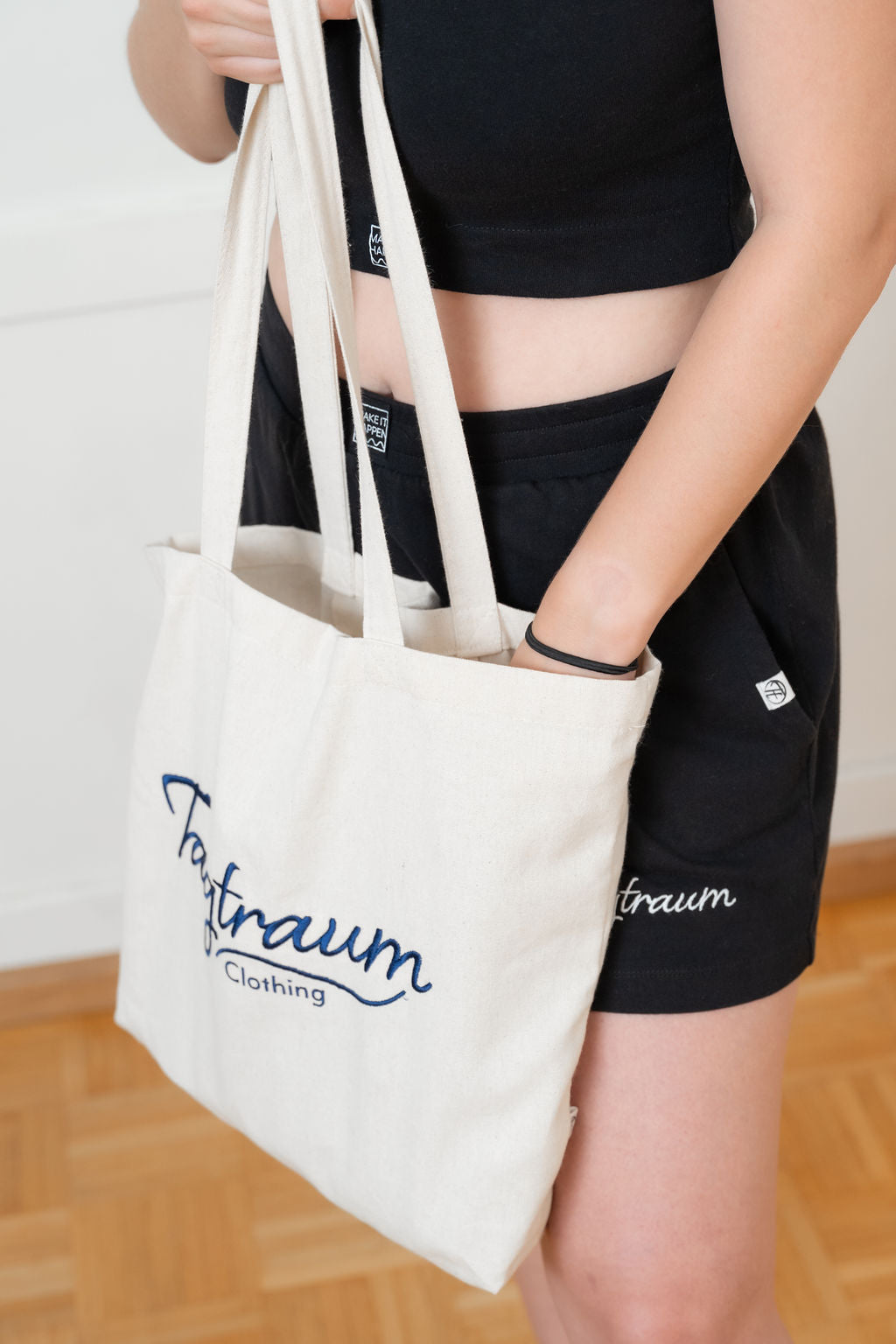 Ein Shopper aus Bio-Baumwolle, naturfarben, mit edlem, dunkelblauem Tagtraum Clothing Stick, getragen von einer Frau, mit der einen Hand sucht sie etwas in der Tasche, mit der anderen hält sie den Shopper. Sie trägt eine Combo aus Tagtraum Crop Top und Shorts