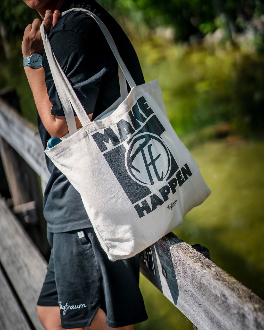 Ein Shopper aus Bio-Baumwolle, naturfarben, mit coolem, schwarzem Tagtraum Print, lässig über die Schulter getragen von einem Mann (Kopf sieht man nicht) , draussen auf einem Steg stehend, eingekleidet in einer Combo aus Tagtraum Shirt und Shorts