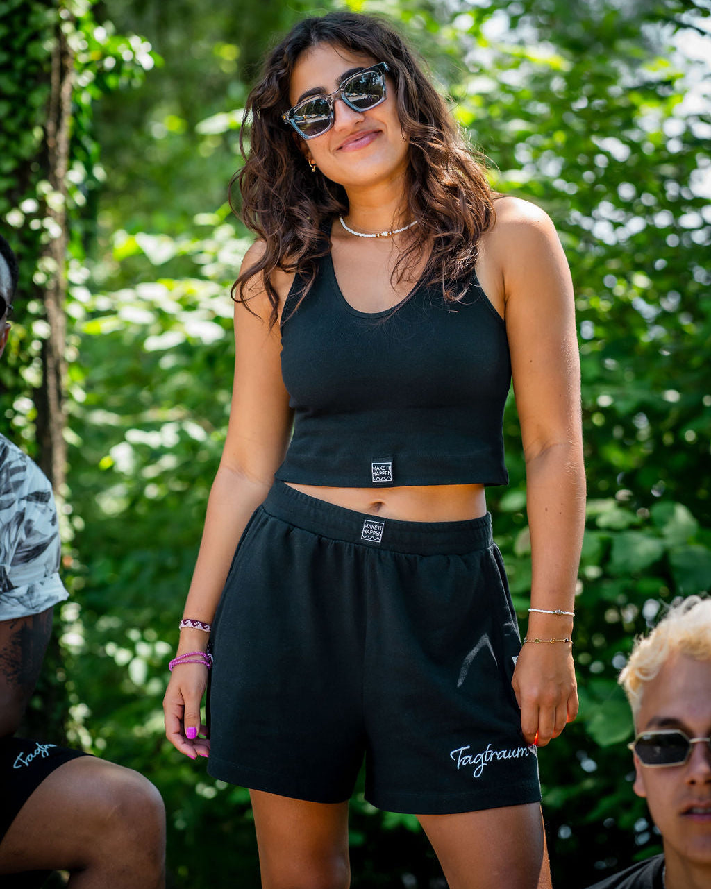 Eine lächelnde, junge Frau mit Sonnenbrille und Locken draussen in der Natur vor grünen Bäumen, mit einem schwarzen Tagtraum Crop Top und einer Shorts, beides aus 100% Bio-Baumwolle