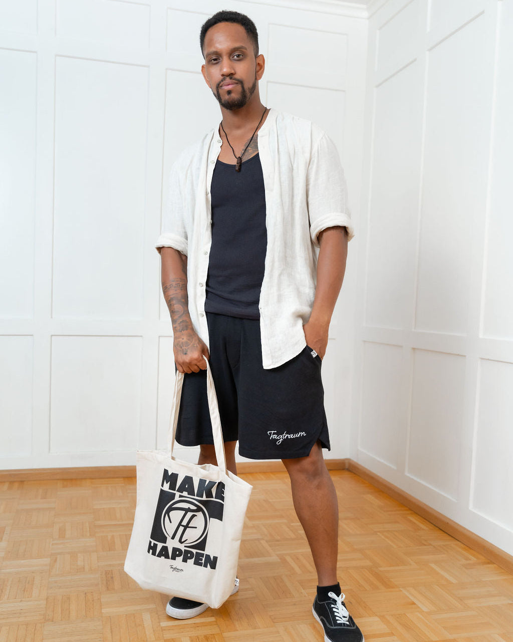 Ein männliches hält den coolen Shopper mit "Make it happen" Print in einer Hand, die andere hat er lässig in der Hosentasche. Er trägt eine schwarze Tagtraum Shorts, ein lässiges Hemd (offen) und darunter ein schwarzes Tanktop