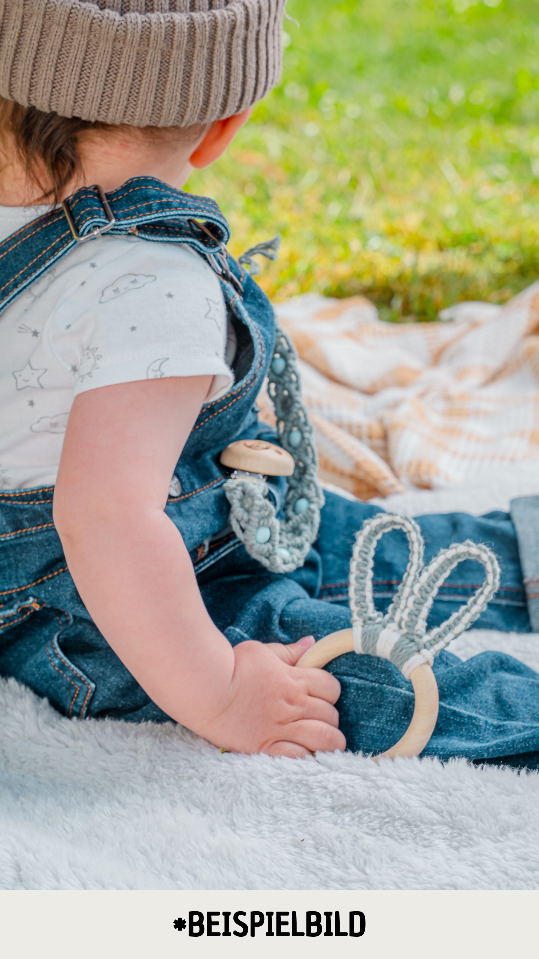 Ein Baby hält einen süssen Hasenohren Beissring in der Hand und sitzt draussen auf einer Pick-Nick Decke, das Gesicht ist nicht zu sehen. Der Beissring ist aus nachhaltigen, umweltfreundlichen Materialien gefertigt und passt perfekt in kleine Baby-Hände. Zudem ist er aus natürlichen Materialien und komplett Schadstofffrei. Handgefertigt in der Schweiz.