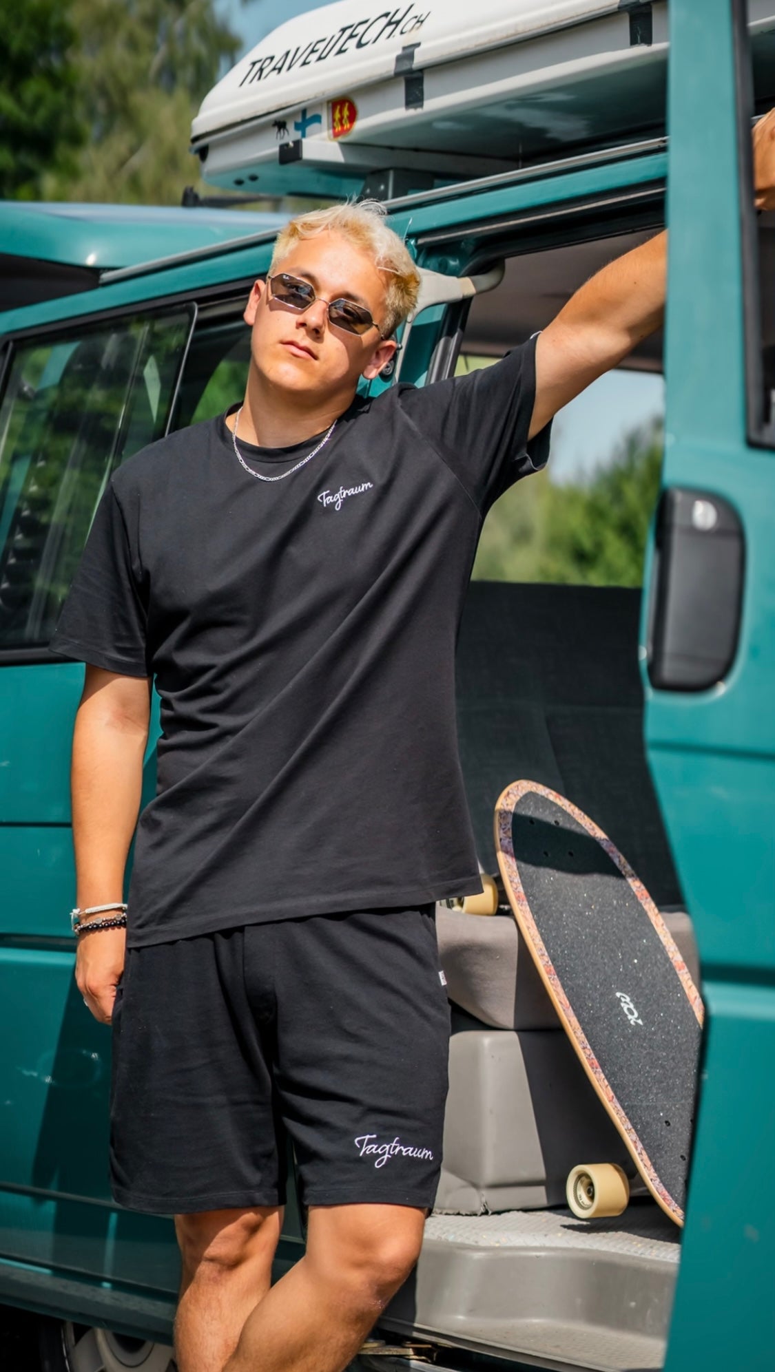 Unser Model, lässig vor dem VW-Bus posierend; mit unserem schwarzen Tagtraum Shirt und unserer ultra bequemen Unisex Shorts; beides aus Bio-Baumwolle. Dazu eine Silbehalskette und eine coole Sonnenbrille; im Hintergrund ein Surfskate.