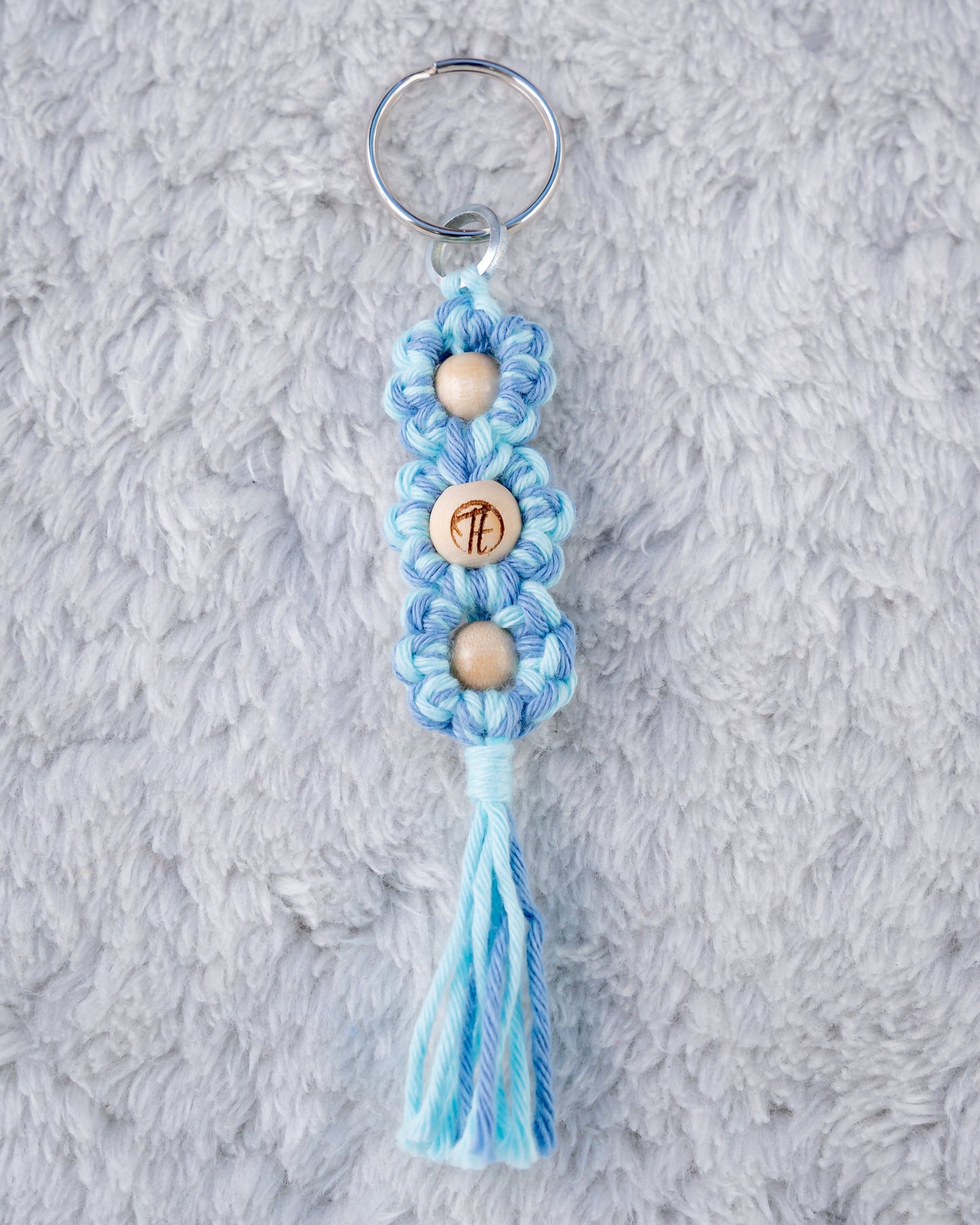 Schöner Makramee Schlüsselanhänger geknüpft aus einem blauen Garnmix, mit drei naturfarbenen Holzperlen, eine davon versehen mit unserem Tagtraum Logo, die anderen Lila und Babyblau. Umweltfreundlich, nachhaltig und handgefertigt von uns in der Schweiz.