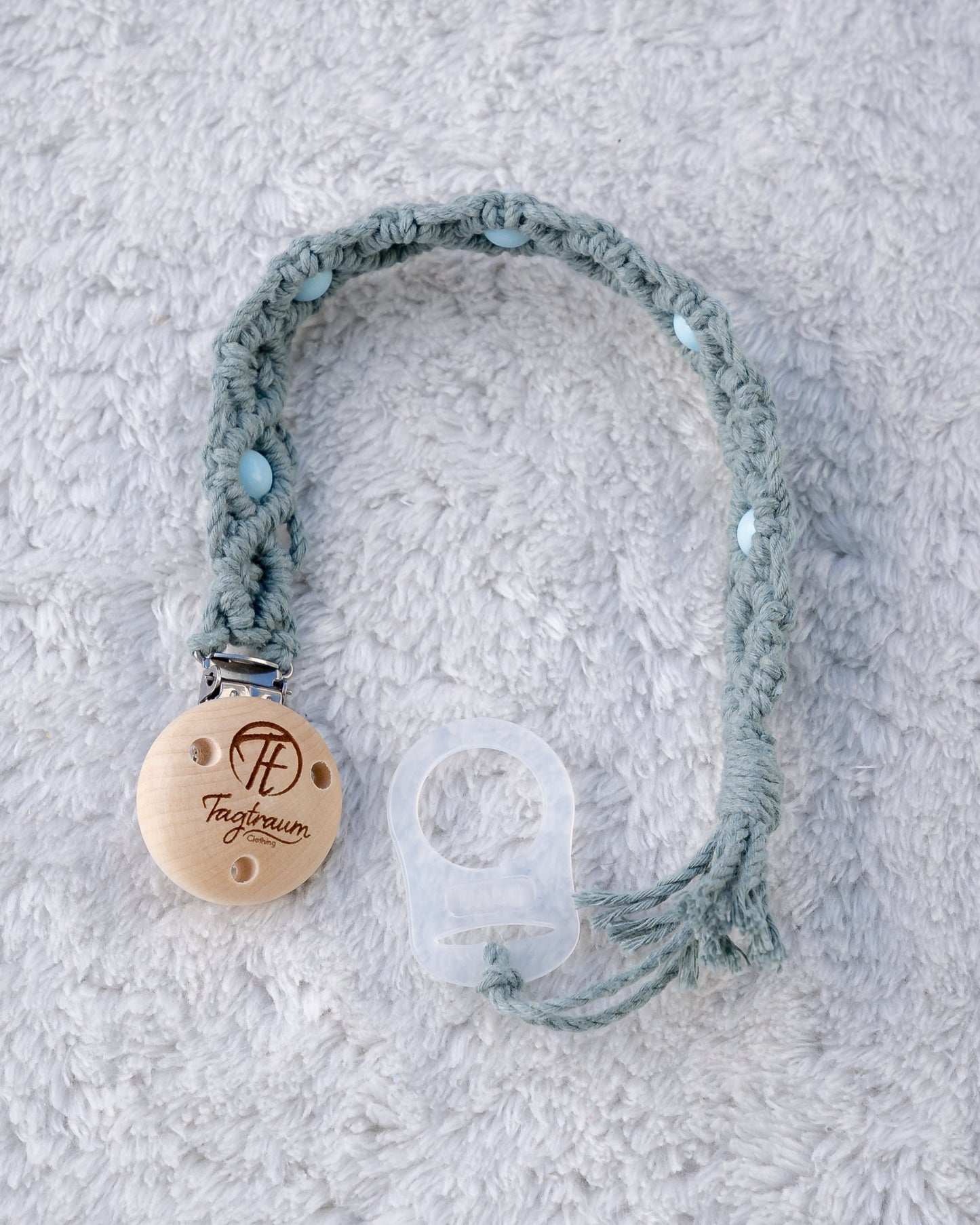 Schöne Schnullerkette aus eukalyptusgrünem Garn mit babyblauen Holzperlen, einem hochwertigen Schnullerclip aus Holz, und einem Adapter für MAM Schnuller. Auf dem Holzclip befindet sich eine dle Tagtraum Cothing Gravur mit Logo. Die Schnullerketten werden handgefertigt von Tagtraum Clothing in der Schweiz.