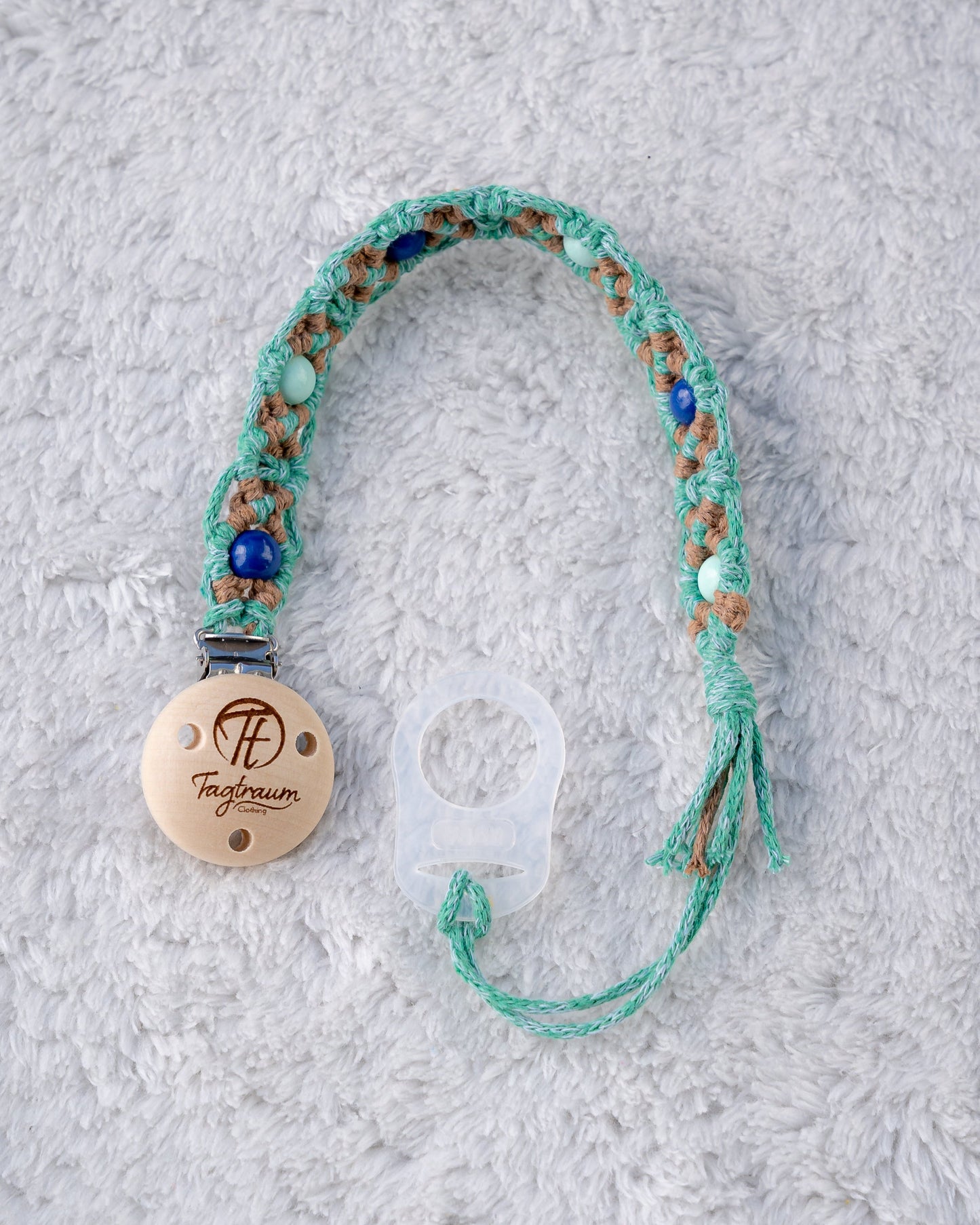 Schöne Schnullerkette aus grünem und braunem Garn mit türkisen und dunkelblauen Holzperlen, einem hochwertigen Schnullerclip aus Holz, und einem Adapter für MAM Schnuller. Auf dem Holzclip befindet sich eine dle Tagtraum Cothing Gravur mit Logo. Die Schnullerketten werden handgefertigt von Tagtraum Clothing in der Schweiz.