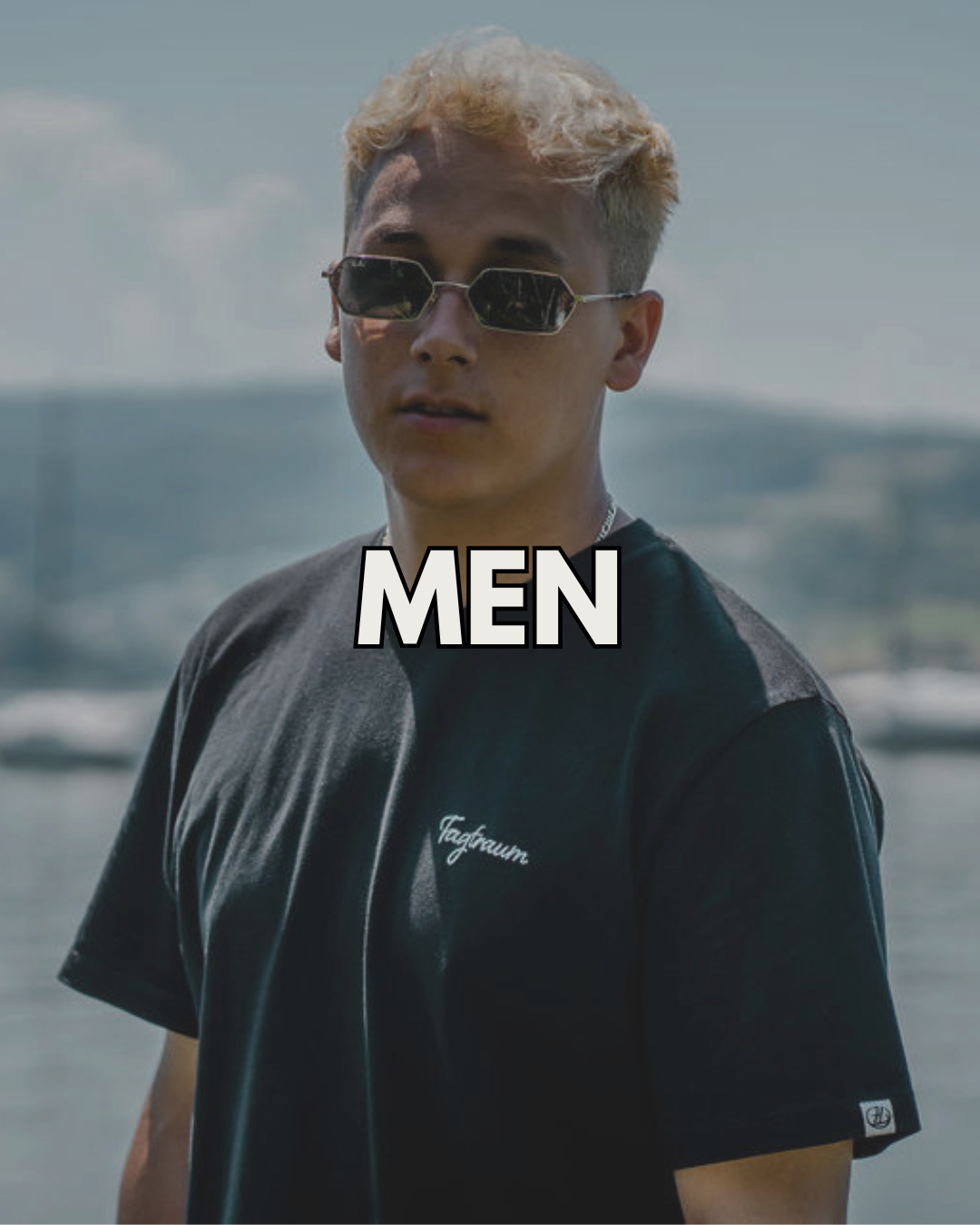 Ein bärtiger Mann sitzt vor einem Weihnachtsbaum, draussen im Schnee, er trägt ein schwarzes Tagtraum Beanie und ein Unisex Shirt, sowie eine blaue Skihose, in der Hand hält er einen Becher mit Tee