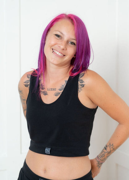 Eine lächelnde junge Frau mit pinken Haaren, Tattoos und einem Lippenpiercing, mit einem schwarzen Tagtraum Crop Top und einer Shorts aus 100% Bio-Baumwolle