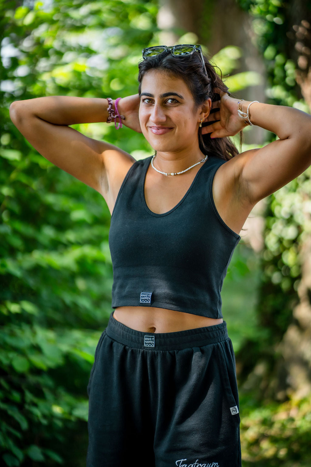 Eine junge Frau mit Sonnenbrille in einem schwarzen Tagtraum Crop Top und einer Shorts, beides aus 100% Bio-Baumwolle. Sie steht draussen in der Natur vor saftig grünen Bäumen