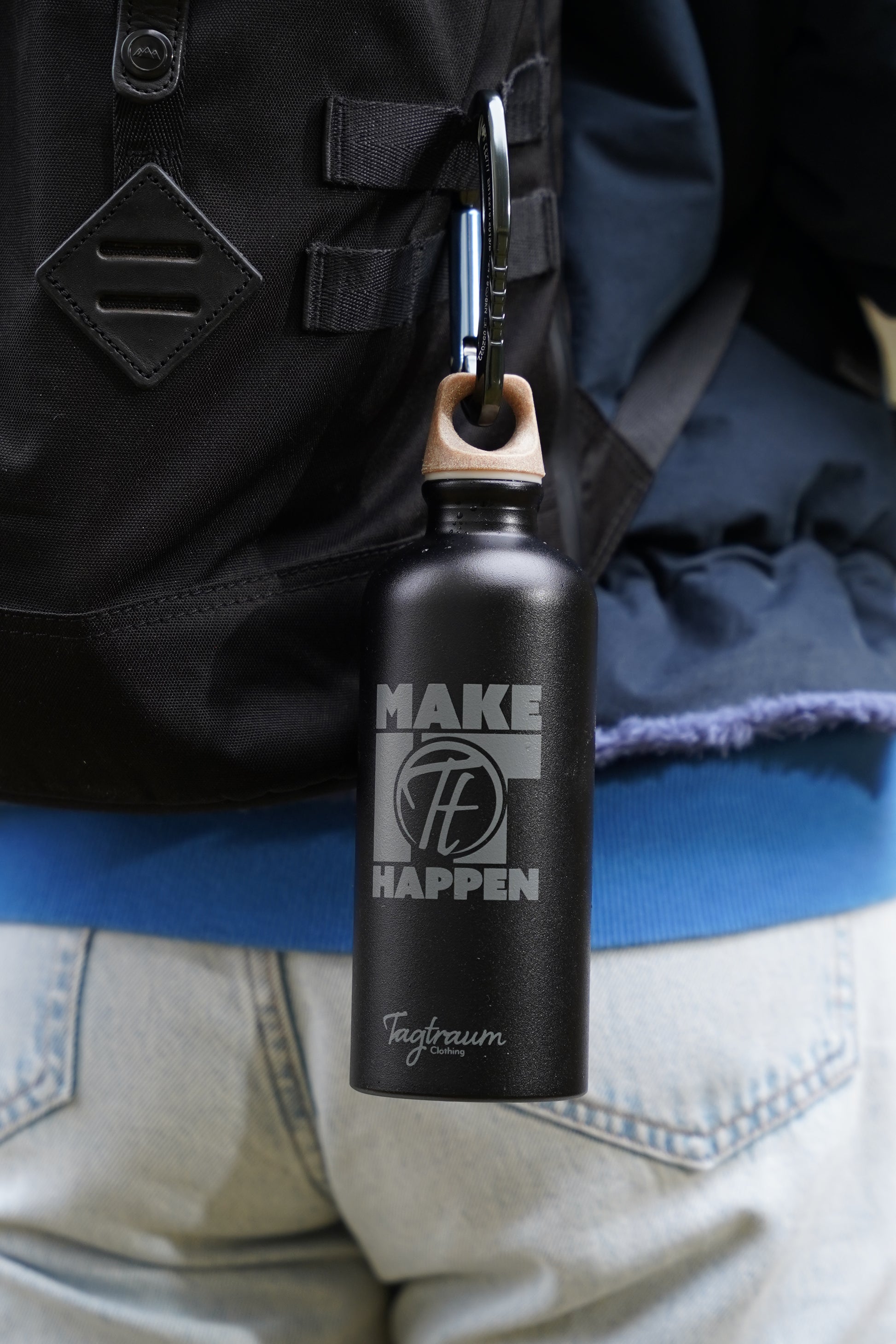 Schwarze Aluminium Trinkflasche aus 100% recycelten Materialien mit einem edlen "Make it happen" Print und einem Verschluss aus Cellulose, angehängt an einem Rucksack, mit einem Karabiner