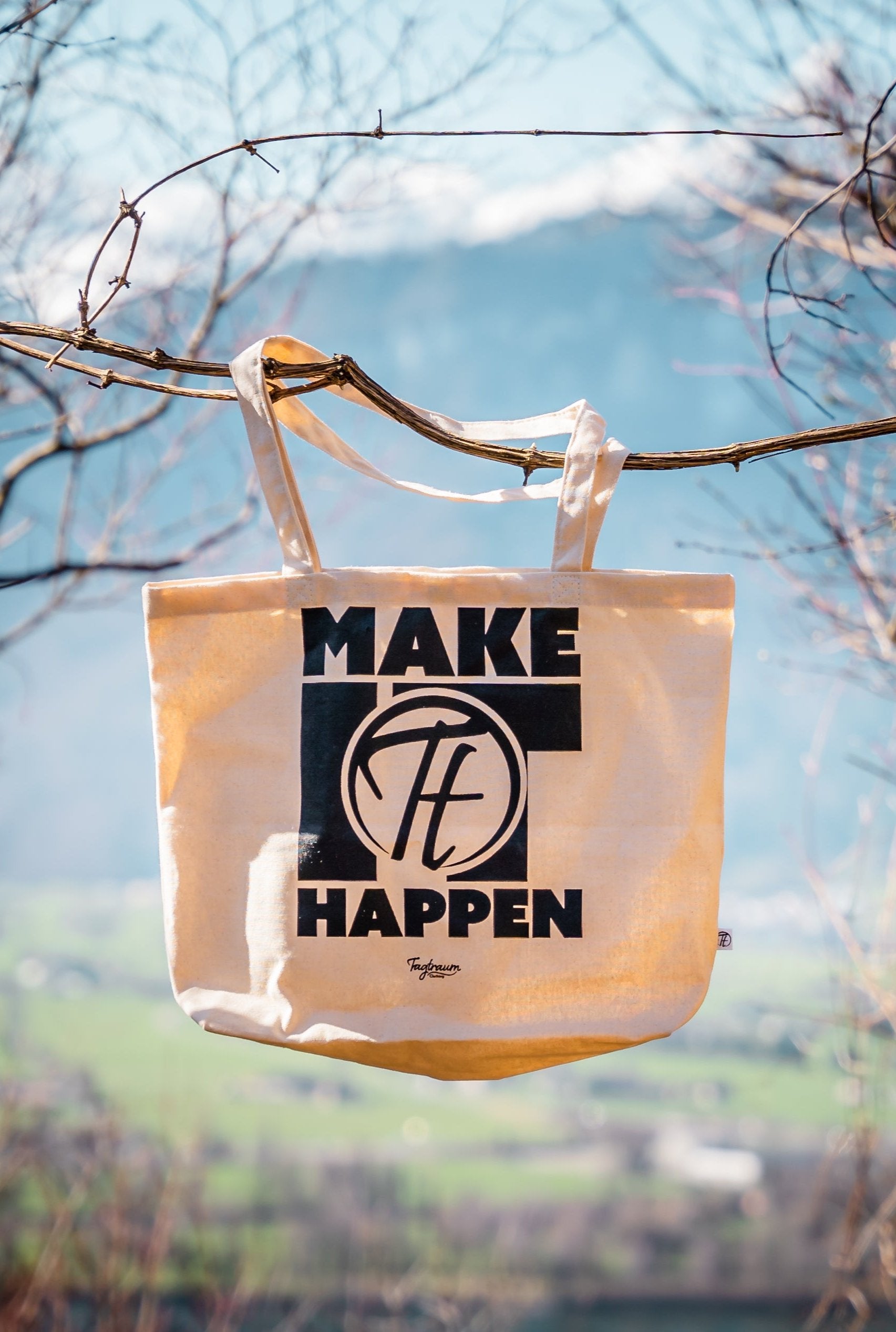 Ein naturfarbener Shopper aus 100% Bio Baumwolle mit einem schwarzen "Make it happen" Print, draussen in Szene gesetzt, an einem Ästchen aufgehängt