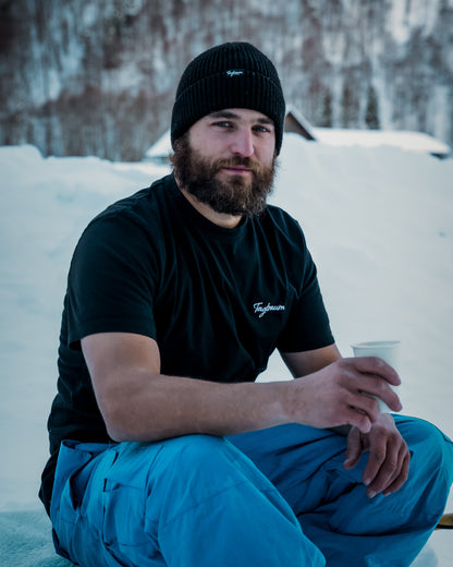 Ein bärtiger Mann mit einem schwarzen Tagtraum Beanie und Unisex Shirt sitzt draussen auf einem Schlitten im Schnee, er hält eine Tasse in der Hand.