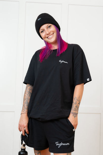 Ein Model mit Tattoos und pinken Haaren lächelt in die Kamera; sie trägt ein schwarzes unisex Bio-Baumwoll Shirt mit edlem weissen Tagtraum Stick auf der linken Brust. Zudem trägt sie ein schwarzes Beanie und Tagtraum Shorts. In der einen Hand hält sie den grossen Tagtraum Karabiner, an dem die Tagtraum Trinkflasche hängt