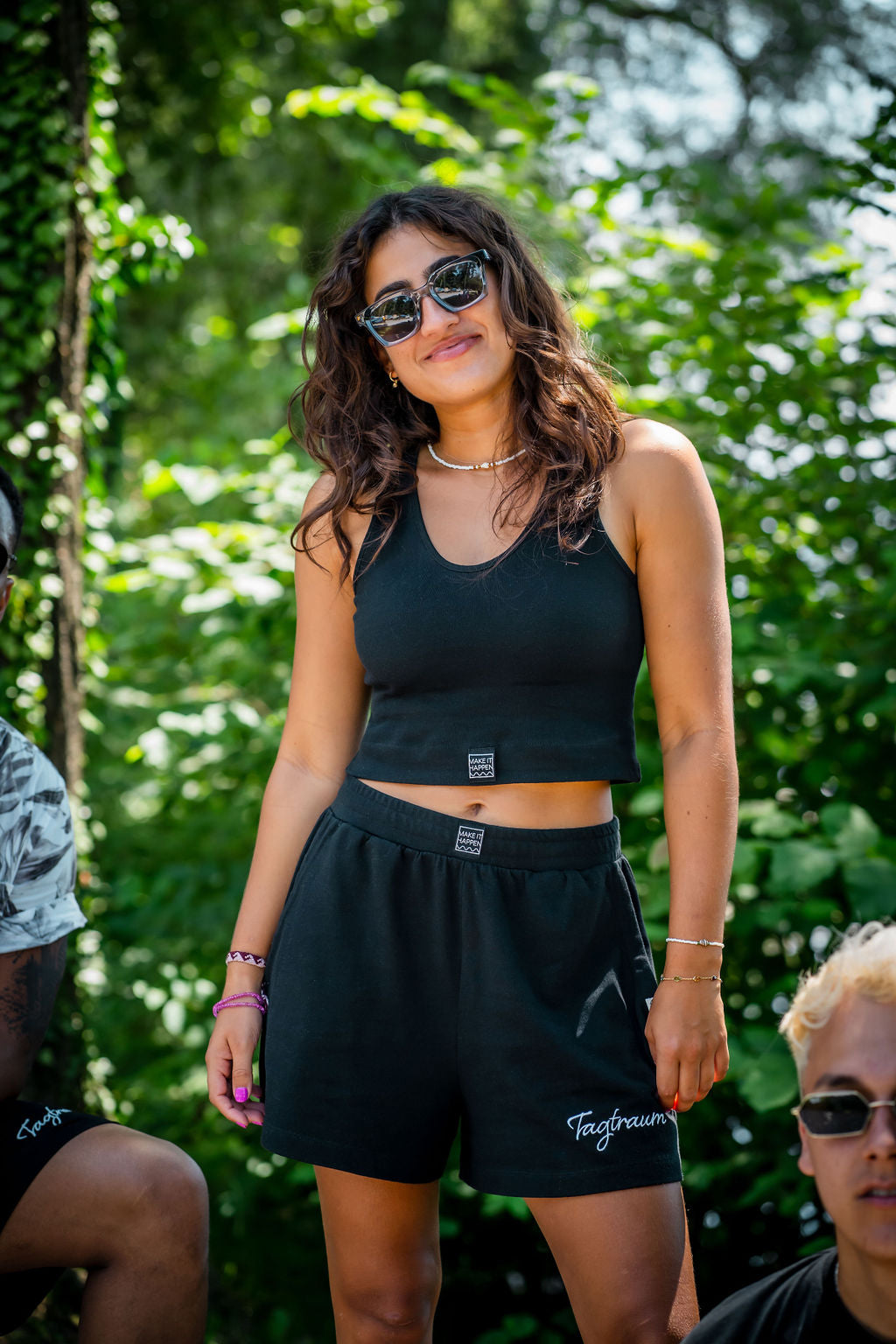 Eine lächelnde, junge Frau mit Sonnenbrille und Locken draussen in der Natur vor grünen Bäumen, mit einem schwarzen Tagtraum Crop Top und einer Shorts, beides aus 100% Bio-Baumwolle