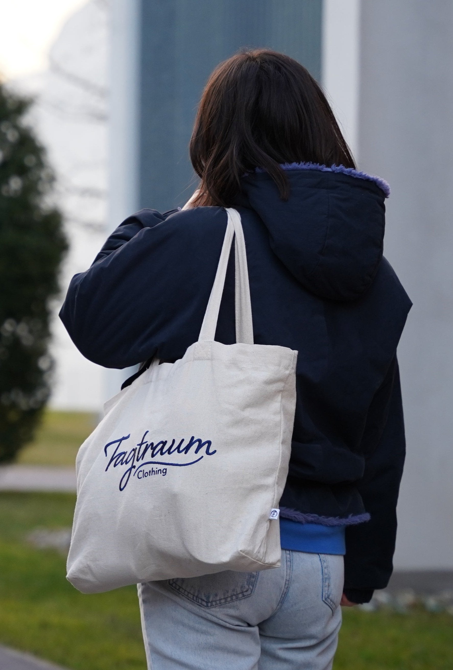 Eine Frau trägt den Baumwoll-Shopper lässig über die linke Schulter. Sie trägt zudem eine dunkelblaue Winterjacke und eine hellblaue Jeans. Man sieht das Model nur von hinten. 