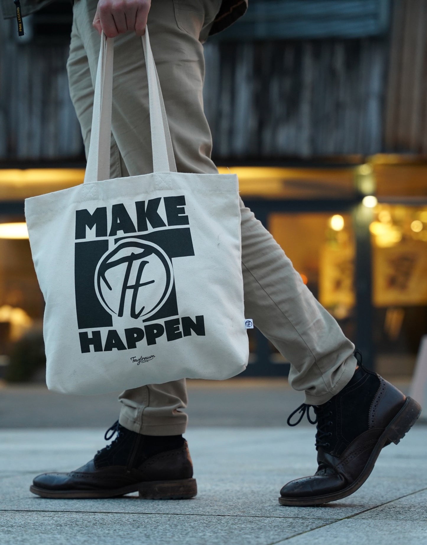 Ein Shopper aus Bio-Baumwolle, naturfarben, mit coolem, schwarzem "Make it happen" Print. Ein Mann hält die Tasche in der Hand und befindet sich draussen in einer urbanen Gegend (man sieht nur seinen Unterkörper). Er trägt eine beige Stoffhose und Winterboots