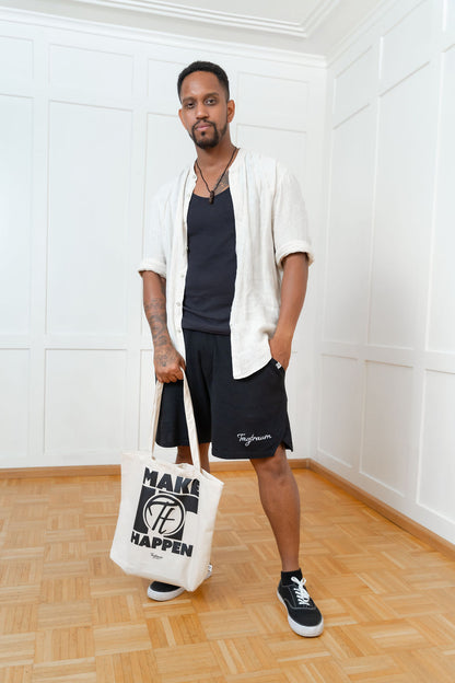 Ein männliches hält den coolen Shopper mit "Make it happen" Print in einer Hand, die andere hat er lässig in der Hosentasche. Er trägt eine schwarze Tagtraum Shorts, ein lässiges Hemd (offen) und darunter ein schwarzes Tanktop