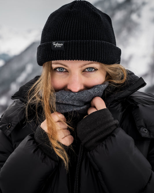 Eine Frau mit einem schwarzen Beanie, blauen Augen, die Jacke über den Mund gezogen, befindet sich draussen, es scheint kalt zu sein