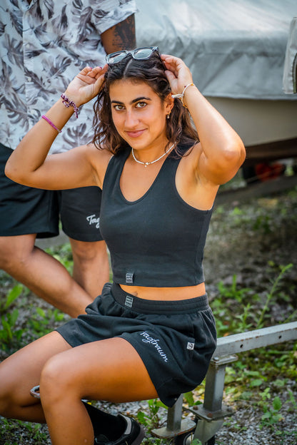 Eine junge Frau mit Sonnenbrille und Locken, sitzend in der Natur, in einem schwarzen Tagtraum Crop Top und einer Shorts, beides aus 100% Bio-Baumwolle