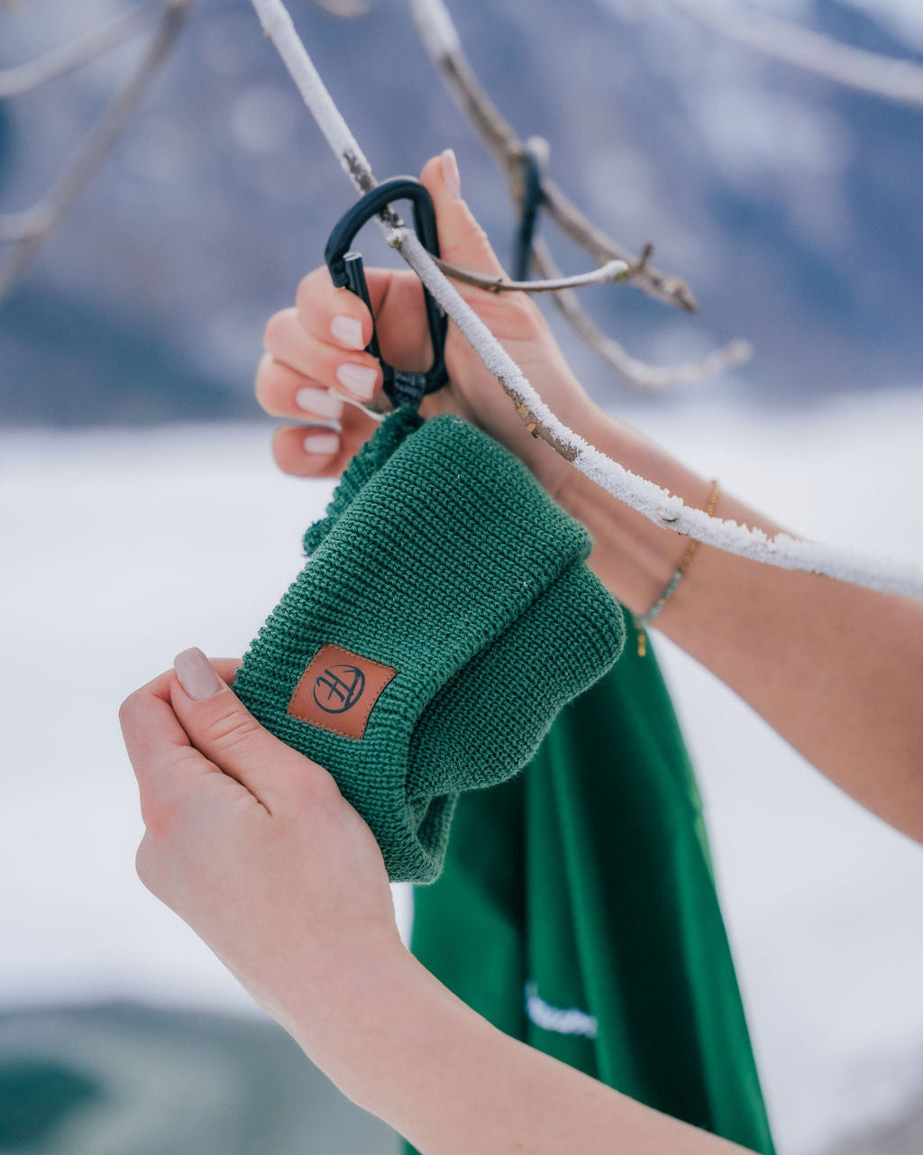 Ein waldgrünes Fisherman Beanie, zwei Hände einer Person, welche das Beanie an einem Larabiner an einen vereisten Ast hängt. Im Hintergrund ein waldgrünes Unisex T-Shirt, ebenfallst aufgehängt aun einem vereisten Ast. Fisherman Beanie und Unisex T-Shirt aus 100% Bio-Baumwolle, Made in Europe. 