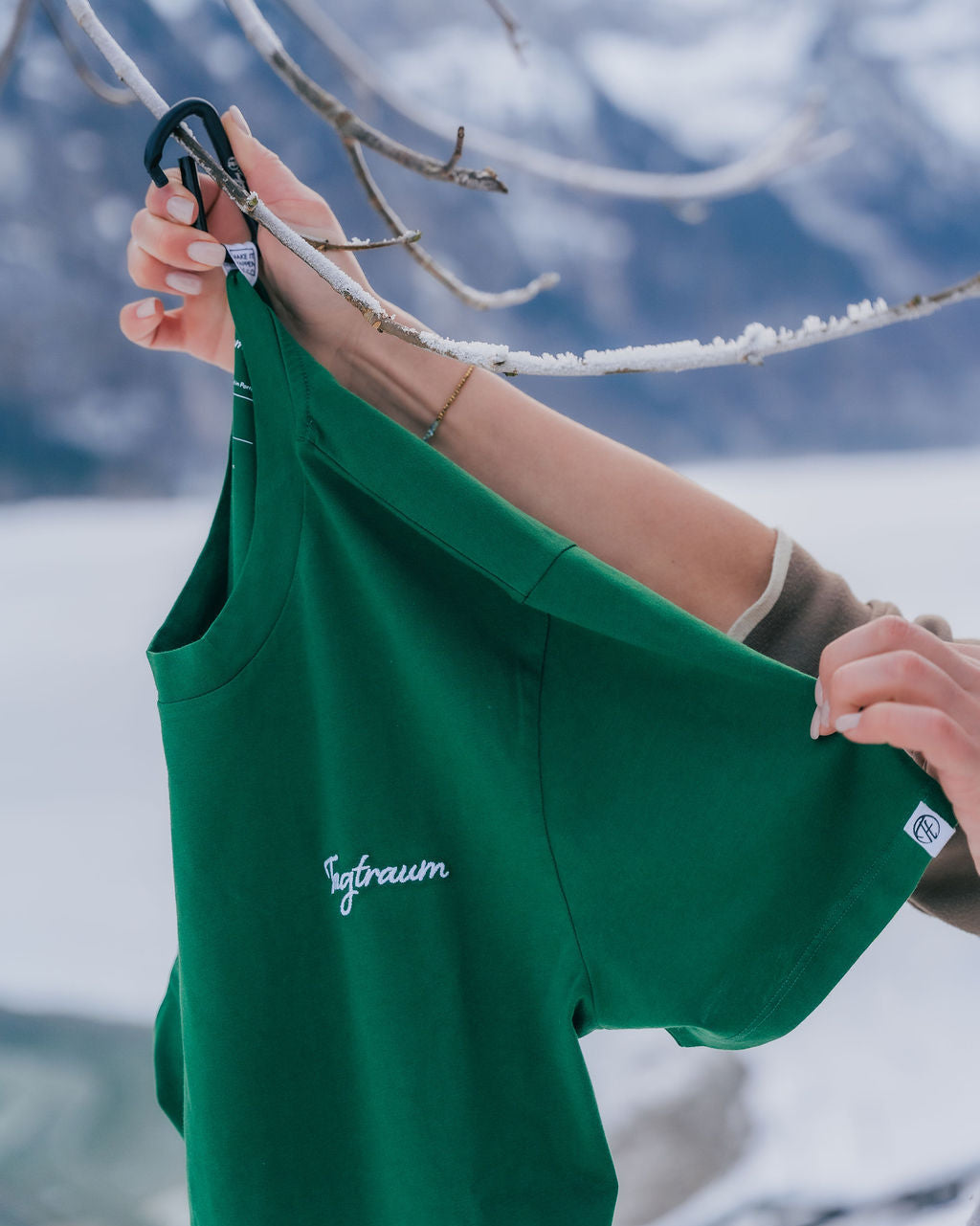 Ein waldgrünes Shirt mit weissem Stick, das von zwei Händen gehalten wird und an einem vereisten Ast in der Natur mit einem Karabiner aufgehängt wird. Tagtraum Clothing Unisex T-Shirt aus 100% Bio-Baumwolle, Made in Europe.