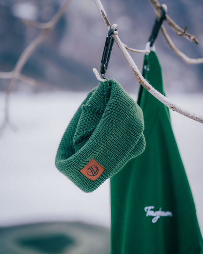 Ein waldgrünes Shirt, sowie ein Fisherman Beanie in derselben Farbe, aufgehängt an vereisten  Ästen in einer Winterlandschaft. Im Hintergrund ein gefrorener See. Fisherman Beanie und Unisex T-Shirt aus 100% Bio-Baumwolle, Made in Europe.