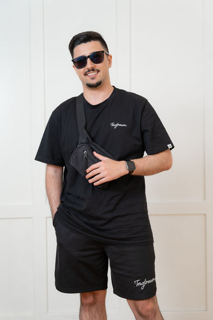 Ein männliches Model in einer schwarzen Tagtraum Shorts und einem schwarzen Unisex T-Shirt, beides aus 100% Bio-Baumwolle und mit einem edlen, weissen Stick versehen. Das Model trägt zudem eine Sonnenbrille und eine Bauchtasche (cross body), er lächels auf dem Foto