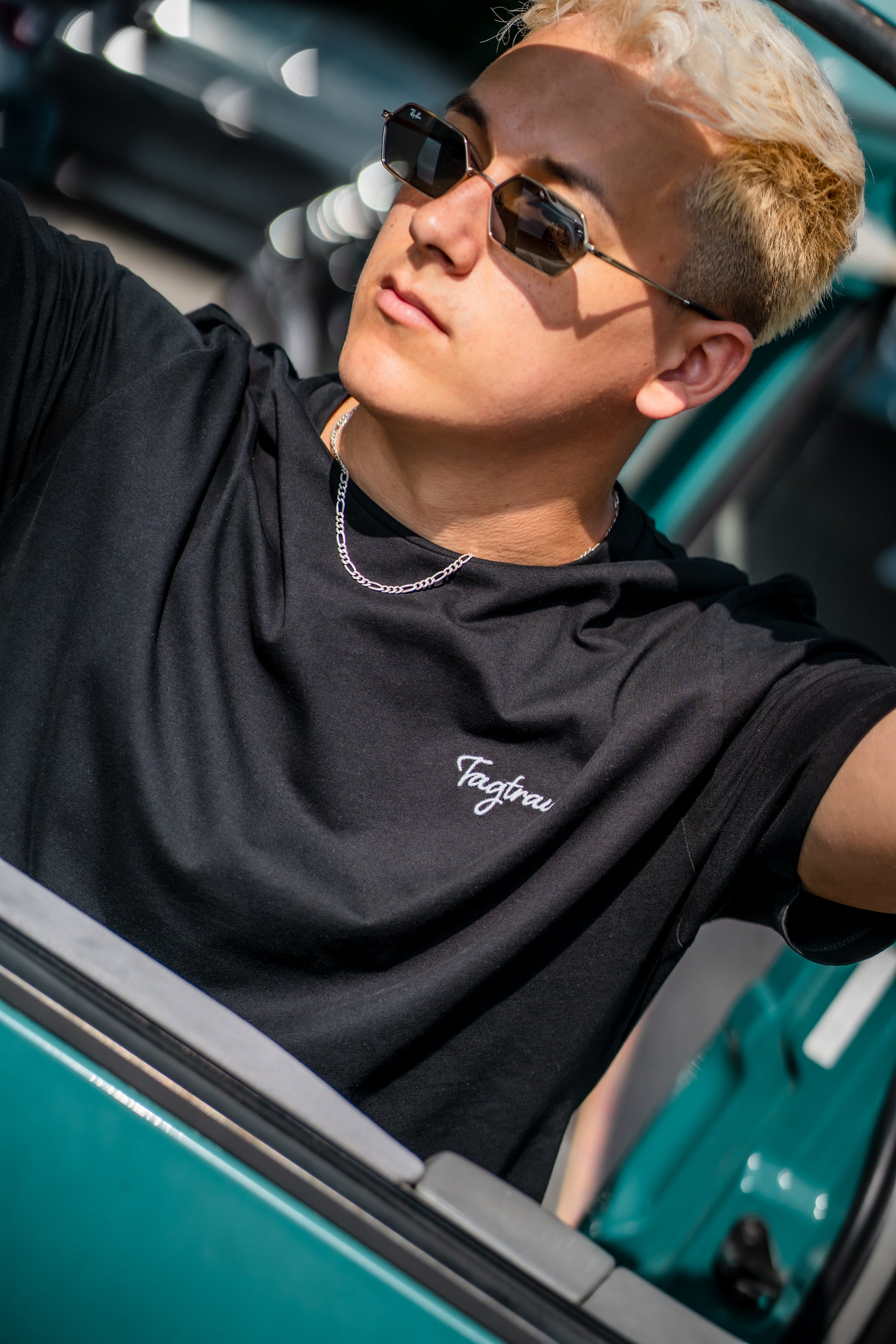 Ein blonder, junger Mann mit cooler Sonnenbrille lehnt sich lässig an die Autotür. Er trägt eine silberne Kette und das schwarze Tagtraum Shirt aus 100% Bio-Baumwolle mit edlem, weissem Stick