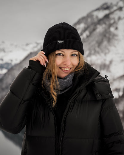 Eine Frau mit blauen Augen lächelt in die Kamera- Sie trägt ein schwarzes Tagtraum Beanie. Sie befindet sich draussen, im Hintergrund verschneite Berge.