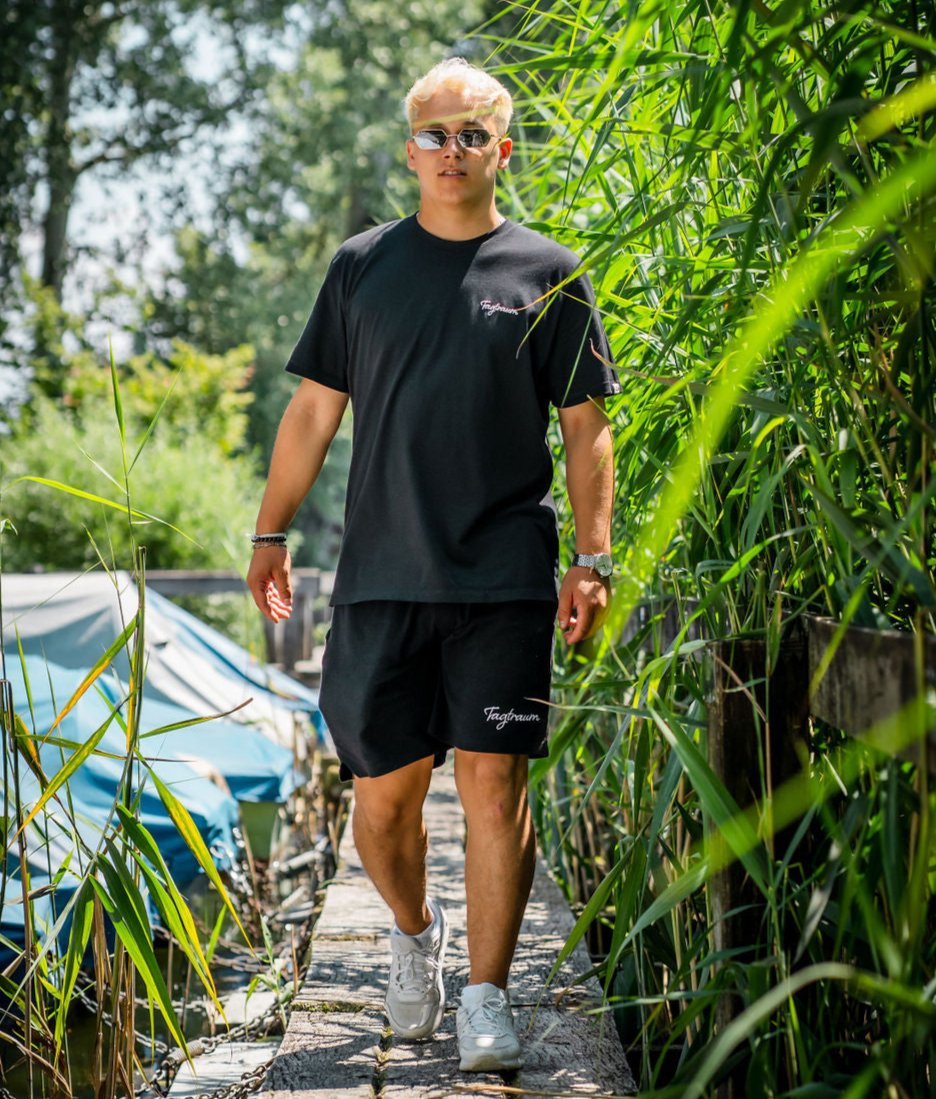 Unser Model in der coolen Kombi aus schwarzem Unisex Shirt und schwarzen unisex Shorts von Tagtraum Clothing; draussen in der Natur, umgeben von Schilf; dazu weisse Sneakers und eine coole Sonnenbrille.