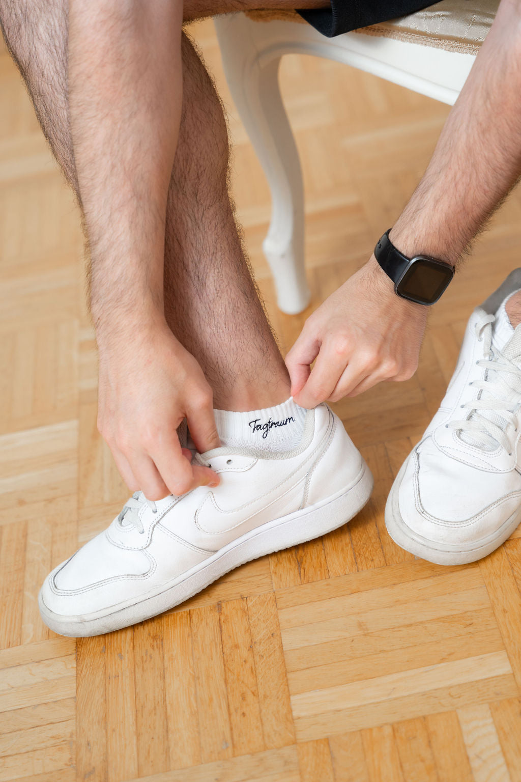 Weisse Sneaker Socken mit einem edlen, schwarzen Tagtraum Stick. Ein männliches Model (nur Beine zu sehen) trägt die Socken und rückt sie mit den Händen zurecht. Zudem trägt er eine Smart Watch und weisse Sneaker