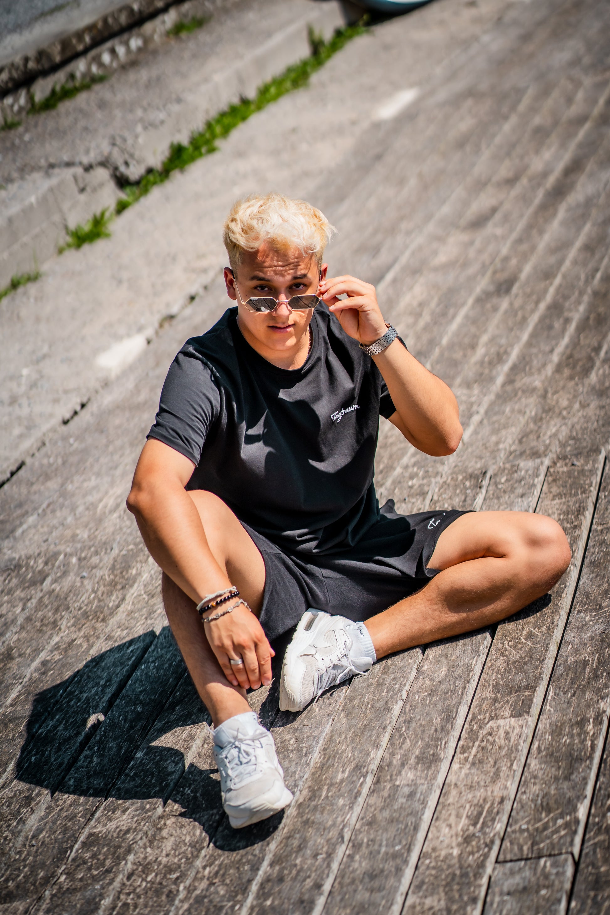 Unser Model in unserem coolen Streetwear Set aus Shirt und Shorts in Schwarz. Schlicht, cool, basic, bequem, nachhaltig. Posierend auf dem Asphalt in der Sonne, mit weissen Sneakern und einer lässigen Sonnenbrille, sowie einer silbernen Uhr. Blick in die Kamera