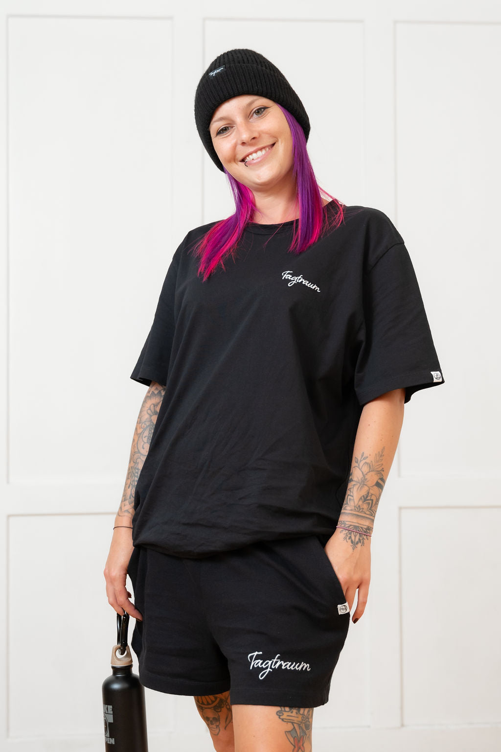 Unser Model in unserem Sommerliebe Set, bestehend aus dem Tagtraum Round Neck unisex Shirt in schwarz, mit edlem Stick und der Frauen Shorts, ebenfalls schwarz, mit weissem Stick. Dazu trägt unser Model ein cooles, schwarzes Beanie und lächelt in die Kamera. Das Shirt lässig-locker getragen.