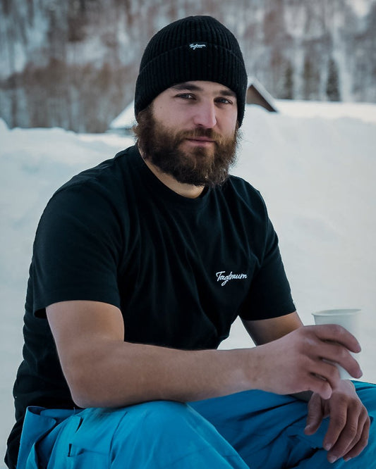 Bärtiger Mann mit schwarzem Tagtraum Shirt und Beanie sitzt in einer Schneelandschaft