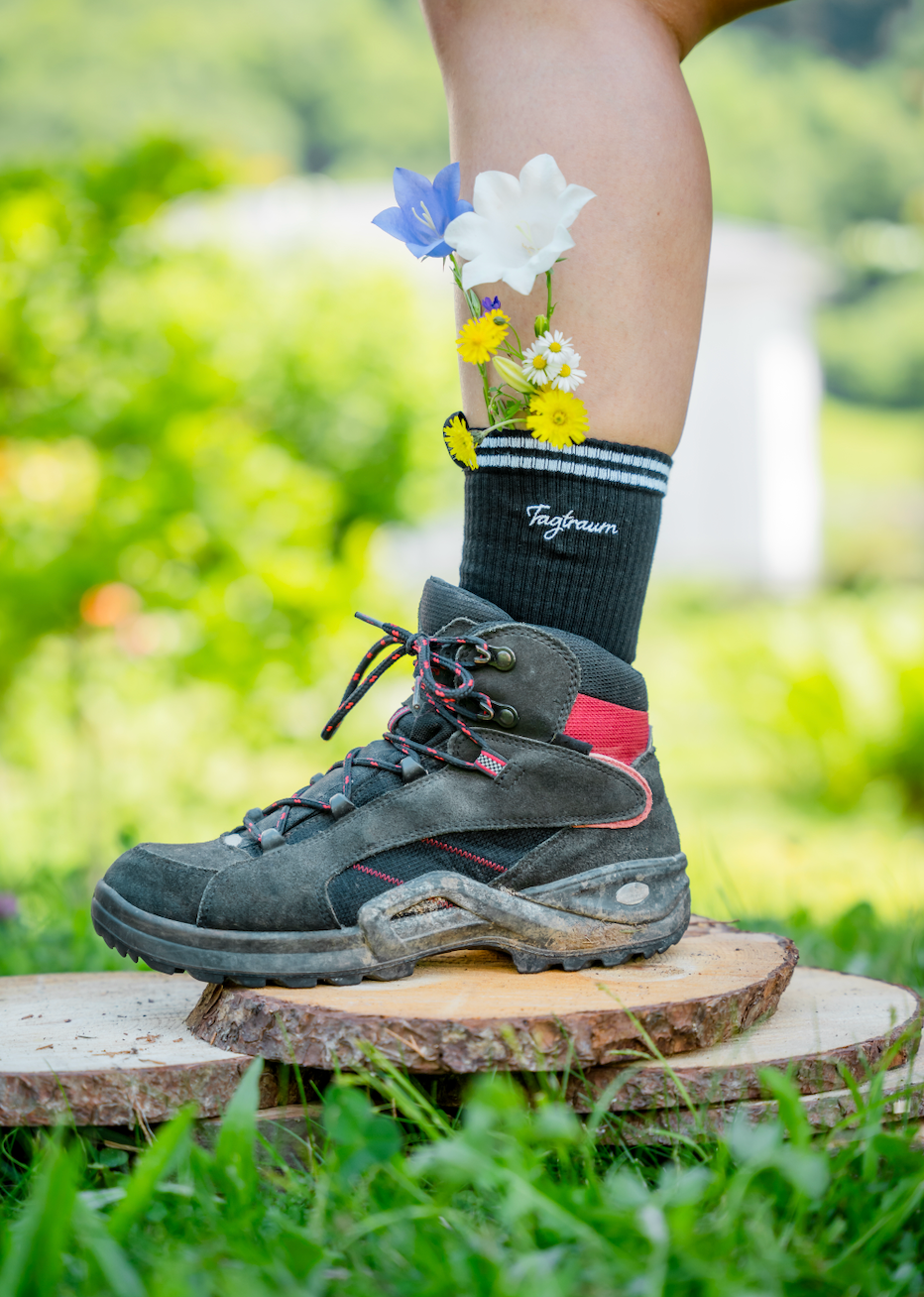 Schwarze Tennissocken mit edlem Tagtraum Stick, getragen in Wanderschuhen, draussen im Grünen. In der Socke stecken ein paar süsse, farbige Wildblümchen