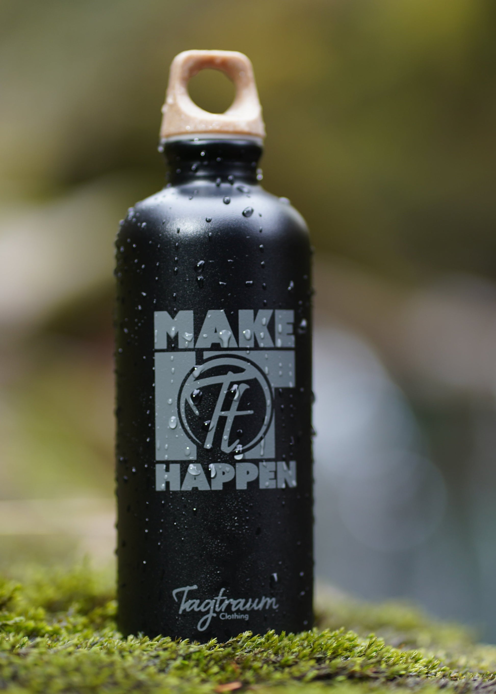 Schwarze Aluminium Trinkflasche aus 100% recycelten Materialien mit einem edlen "Make it happen" Print und einem Verschluss aus Cellulose, fotografiert in der Natur mit Wassertropfen auf der Flasche und auf schönem Moos 