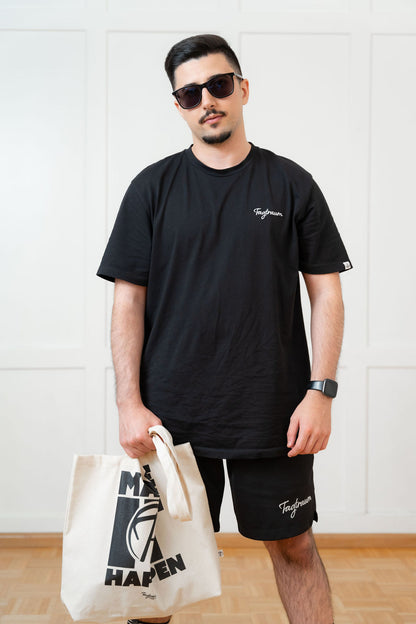 Ein männliches Model mit Sonnenbrille steht frontal zur Kamera und trägt lässig ein schwarzes Bio-Baumwoll Tagtraum Shirt, dazu die Tagtraum Shorts, eine Smartwatch und in einer Hand hält er cool den "Make it happen" Shopper. Das Bild vermittelt Streetstyle pur