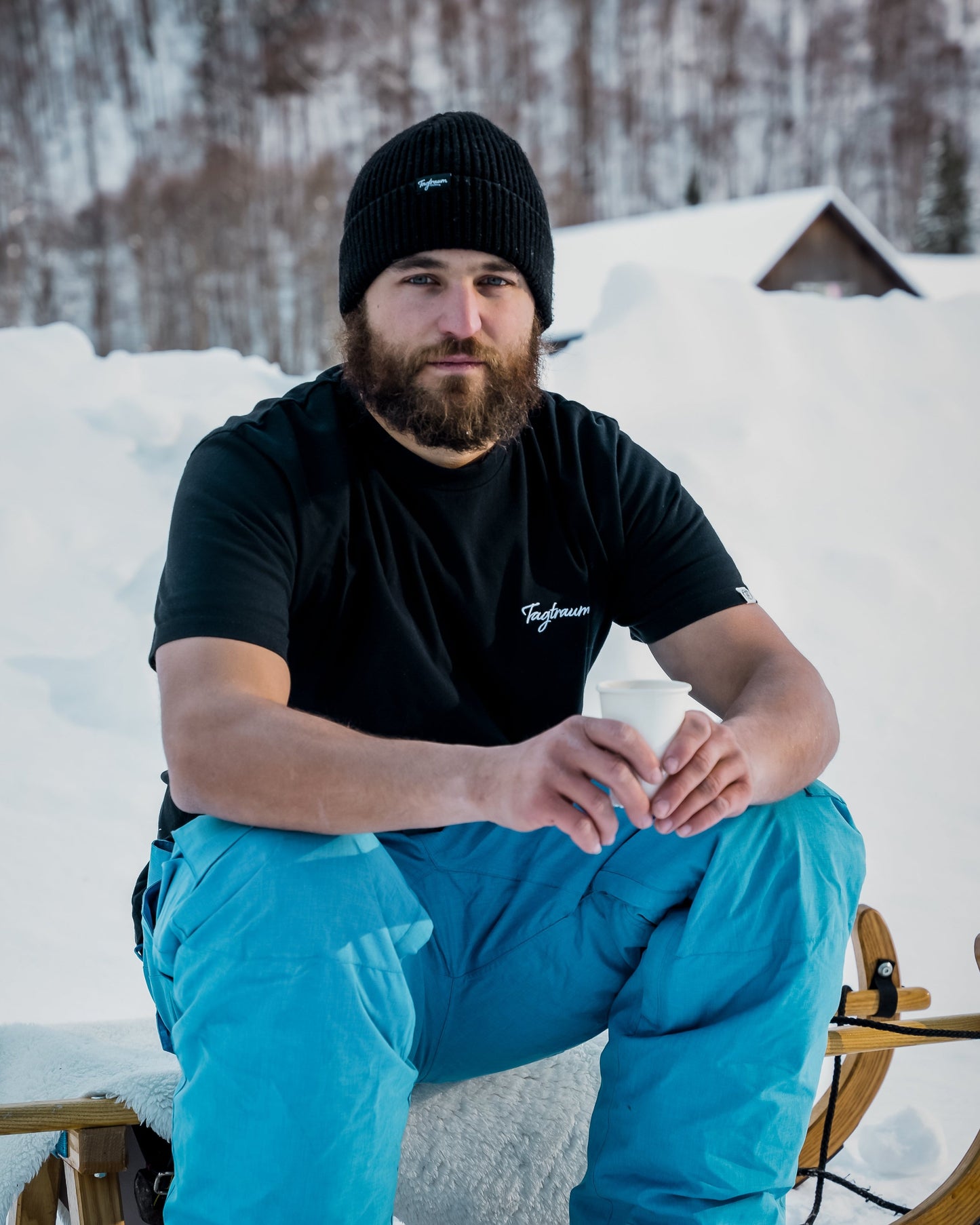 Ein bärtiger Mann sitzt draussen im Schnee auf einem Schlitten. Er schaut in die Kamera, trägt ein schwarzes Tatraum Beanie, ein Unisex Shirt von Tagtraum und eine blaue Skihose, sowie Skischuhe. In der Hand hält er einen Becher mit Tee.